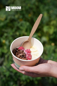 Reusable biodegradable spoon in hand, serving dessert in a cup with raspberries and granola, outdoors.