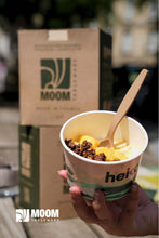 Load image into Gallery viewer, Person holding a cup of dessert with a wooden spoon, featuring MOOM packaging in the background, emphasizing sustainable tableware.