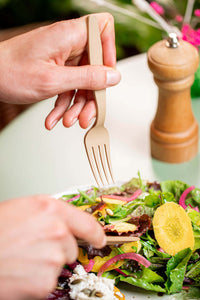 Main hand holding a reusable 16cm fork, picking salad from a plate, with a wooden pepper grinder in background.