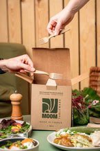 Laden Sie das Bild in den Galerie-Viewer, Two hands placing reusable 16cm forks into a kraft box labeled &quot;MOOM Made in France&quot; with food items on the table.